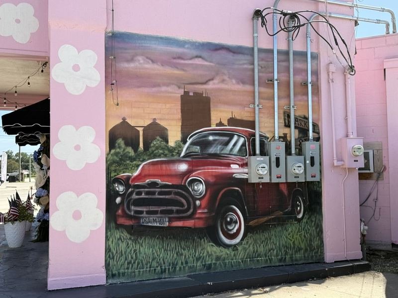Photorealistic mural of a red 1950s pickup truck in tall grass with the ALICO building and silos on the skyline, painted on a pink flower shop wall